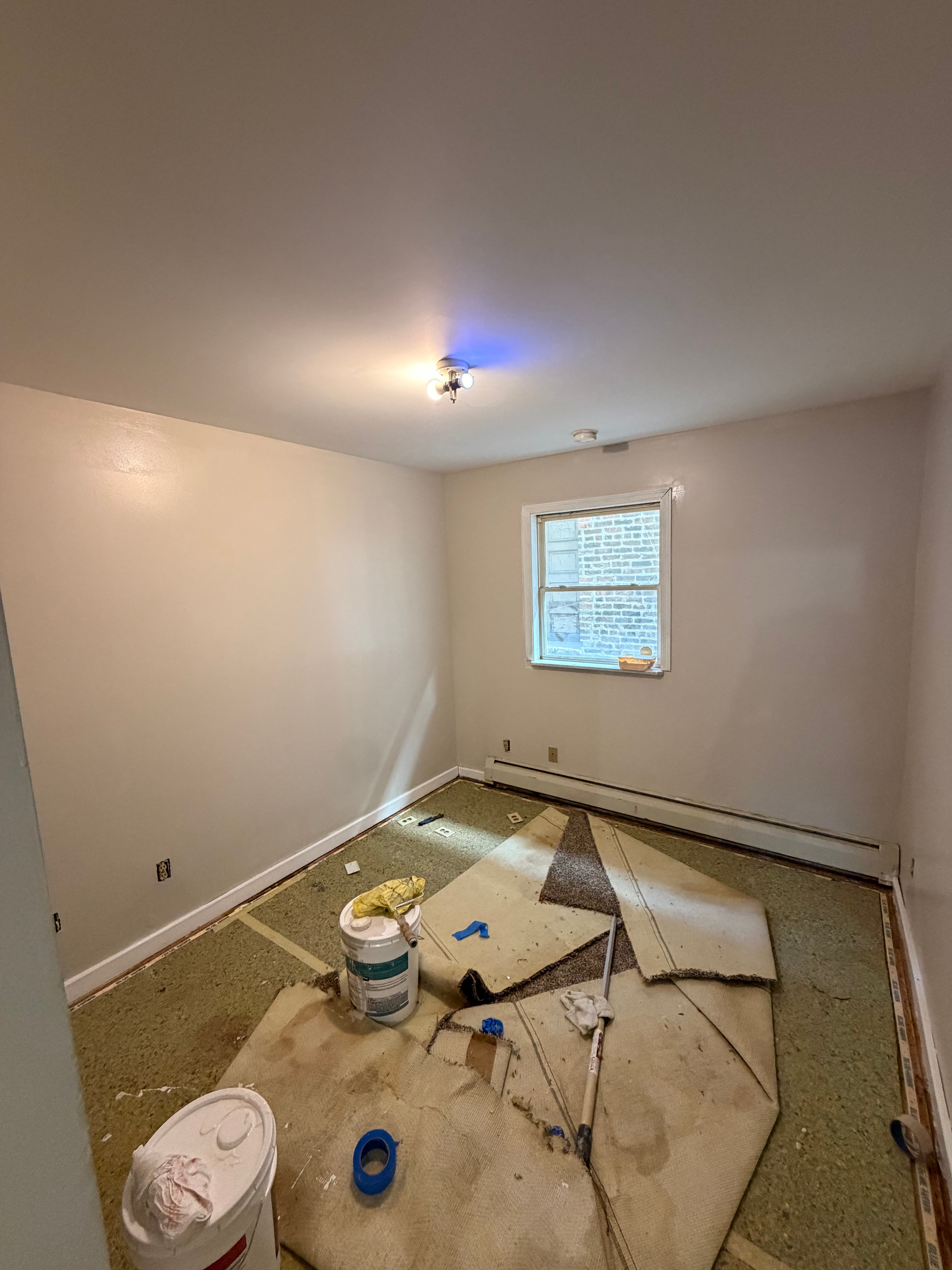 Empty room undergoing renovation with beige walls, pulled-up carpet, and painting supplies on the floor.