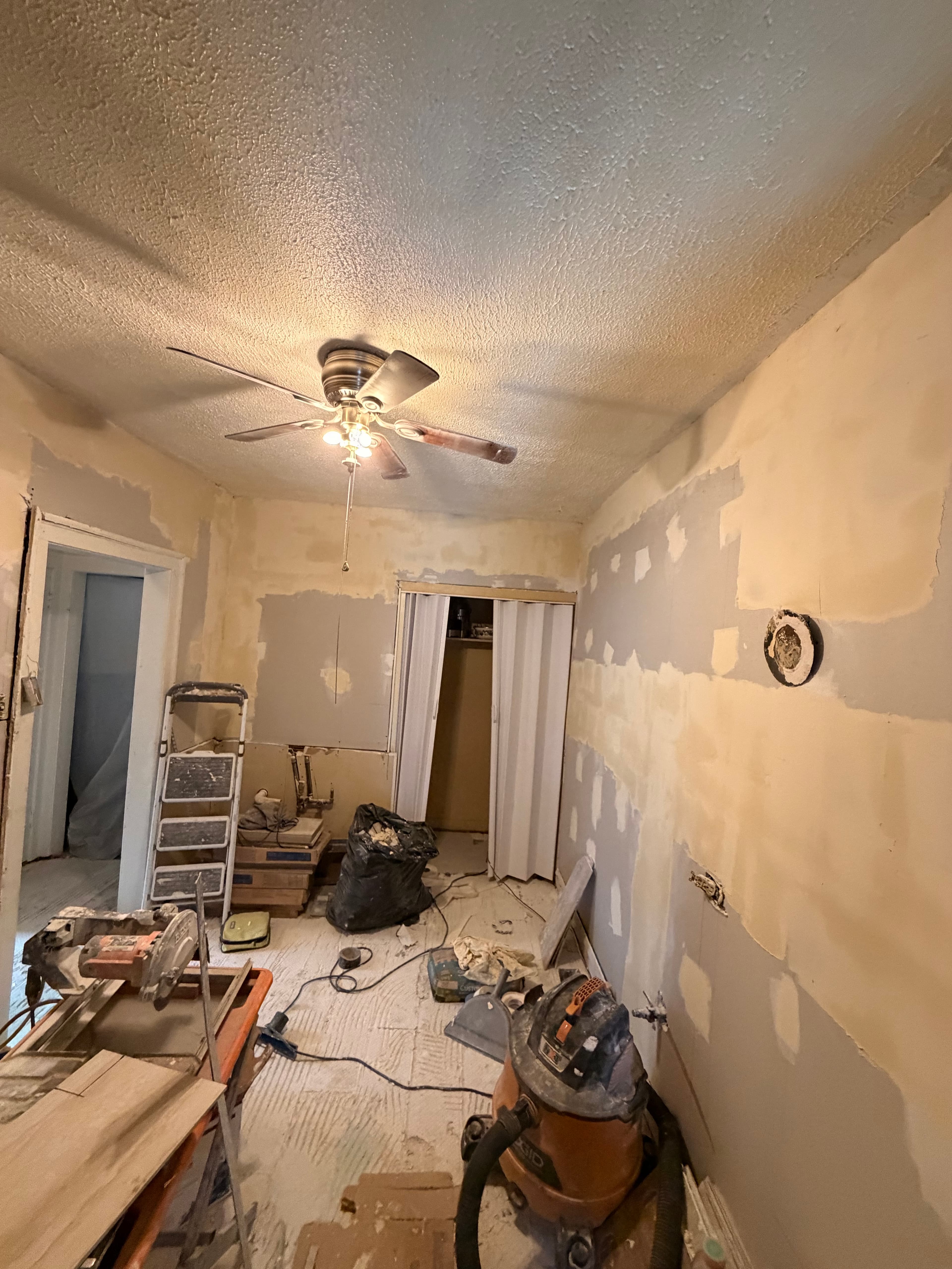 Messy room during renovation featuring patched walls, construction tools, and a lit ceiling fan.